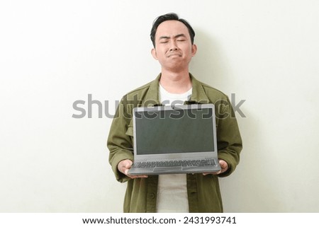 portrait of asian man holding and showing laptop screen with sad face. Indonesian man in green shirt on isolated white background. illustration of a businessman, entrepreneur, freelancer or student