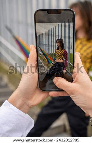woman's hands holding a smartphone taking a photo of a young man with an umbrella in the colors of pride.