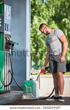 Handsome driver refueling canister with gasoline. Man filling cistern with fuel in case of unforeseen circumstances. Man in casual clothes with pump nozzle on background of modern gas station. Royalty-Free Stock Photo #2432019107