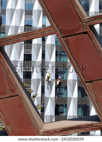 This high-resolution stock photo captures the dynamic interaction between human activity and urban architecture, featuring window washers suspended on an abstract, geometric building facade.