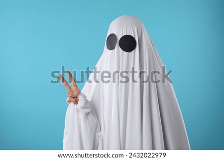 Funny ghost. Woman covered with white sheet showing V-sign on light blue background