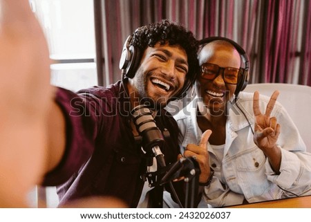 Cheerful podcasters hosts in studio together. A man and woman are seen sharing a joyful moment, laughing and gesturing peace sign while taking a selfie.