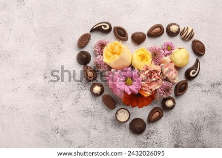 Heart made with delicious chocolate candies and beautiful flowers on grey table, flat lay. Space for text