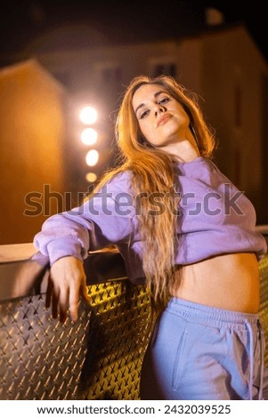 Vertical photo of a blonde beauty trap dancer posing leaning on a urban rail at night