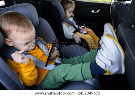 Cute siblings sitting in baby booster car seats and using mobile phones during the road trip. Technology and travel concepts.