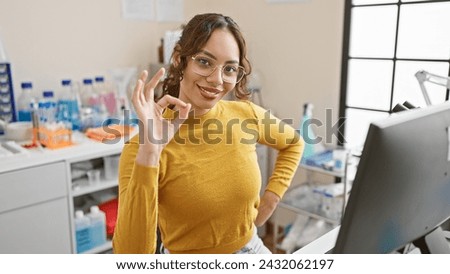 Smiling woman gesturing okay in laboratory with flasks and equipment showing positivity and professionalism.