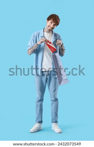 Young tattooed man with gift voucher and shopping bag on blue background