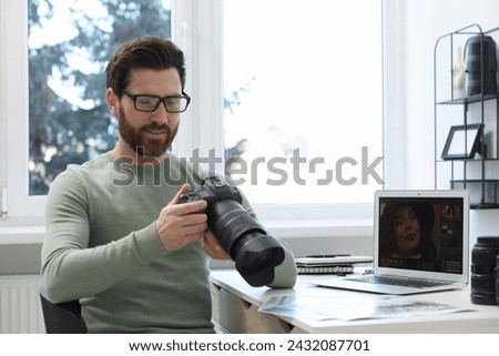 Professional photographer in glasses holding digital camera at table in office, space for text