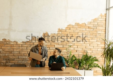 Two smiling professionals engage in a collaborative work session at a wooden table, their camaraderie evident in a contemporary office setting with an exposed brick wall backdrop Royalty-Free Stock Photo #2432101379