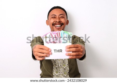 An excited Indonesian Muslim man in koko and peci holds a white envelope labeled THR, filled with money, representing his Religious Holiday Allowance. Isolated on a white background