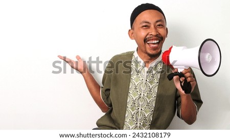 Excited Indonesian Muslim man in koko and peci gestures to the side with an open hand, holding a megaphone. He directs attention to empty copy space for text or advertisements. Isolated on a White