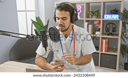 Handsome hispanic man in medical uniform podcasting with microphone and smartphone in radio studio.