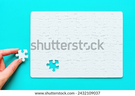 Top view of a hand placing the final piece of a blank rectangular jigsaw puzzle set on a pristine blue background. Royalty-Free Stock Photo #2432109037