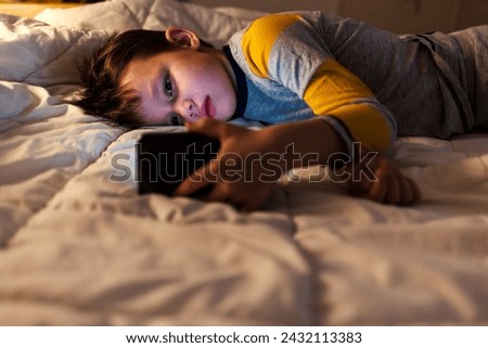 Young boy laying in bed and watching clips by smartphone. Little boy play cell phone at night.