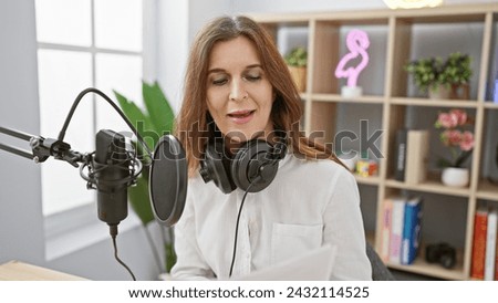 Middle-aged woman recording in podcast studio with microphone and headphones indoors.