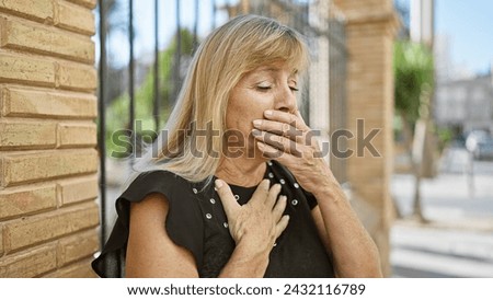 Caucasian blonde, middle age woman looking serious yet relaxed, standing outdoors in sunny city street coughing, showing signs of flu, possibly covid-19, indicating urban infection concern Royalty-Free Stock Photo #2432116789