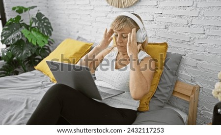 Headache hell, beautiful middle age blonde woman suffering an intense ache, sitting on bed with laptop and headphones in bedroom