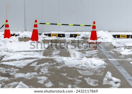 Bright orange traffic cones standing on car park, security barrier with snow. Plastic reflective road cones on snowy winter urban street against cars parking