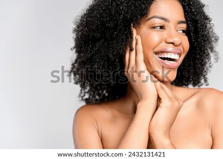 A joyful African-American woman with a vivacious smile and natural curly hair poses with her hands gently framing her face, highlighting her cheerful demeanor and radiant skin against grey background Royalty-Free Stock Photo #2432131421