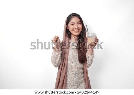 Portrait of an excited young Asian woman raising fist up gesture, winning and celebrating victory pose while holding coffee in a plastic cup. Ramadan and Eid Mubarak concept