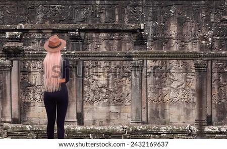 A beautiful blonde woman with a stylish hat wearing black tight trousers watching Bas-relief - Bas-relief at Bayon temple in Angkor Thom. Siem Reap. Cambodia. Panorama