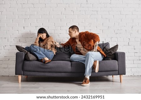 young irritated man talking to young asian wife crying on couch in living room, divorce concept Royalty-Free Stock Photo #2432169951