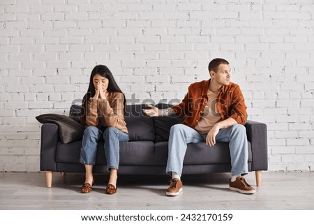 discouraged man pointing at offended asian wife on couch in living room, relationship difficulties Royalty-Free Stock Photo #2432170159