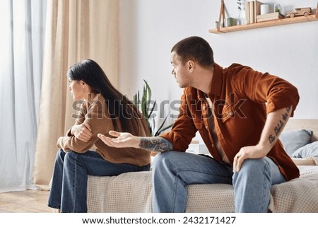 angry tattooed man quarreling at young asian wife white sitting in bedroom at home, divorce concept. Translation of tattoo: be faithful to the one who is faithful to you Royalty-Free Stock Photo #2432171427