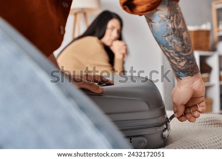 tattooed husband packing suitcase near offended asian wife crying on bed at home, divorce concept Royalty-Free Stock Photo #2432172051