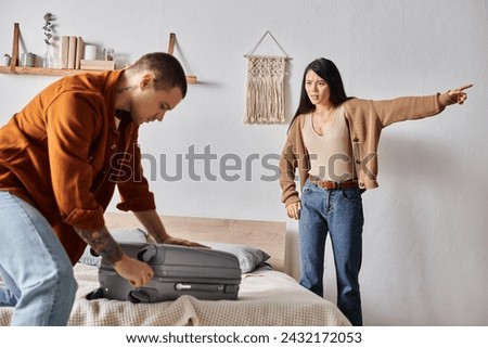 frustrated asian woman pointing away near husband packing suitcase at home, relationship trouble