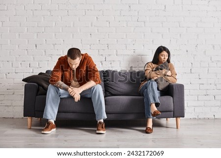 young offended interracial couple sitting on couch in modern living room, family divorce concept Royalty-Free Stock Photo #2432172069