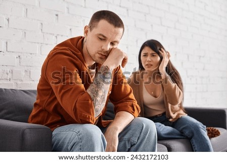 frustrated asian woman looking at sad tattooed husband on couch at home, family divorce concept Royalty-Free Stock Photo #2432172103