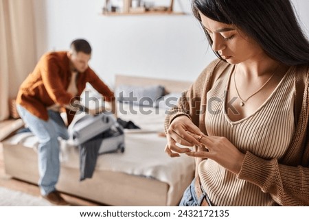 asian woman taking off wedding ring while husband packing suitcase in bedroom, family divorce Royalty-Free Stock Photo #2432172135