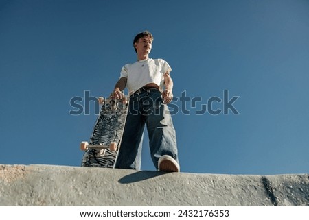 Low angle of happy male skateboarder holding skateboard standing at skate park in sunny day Royalty-Free Stock Photo #2432176353