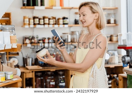 Thoughtful woman using smartphone and buying products at store. Сonscious shopping and retail of organic food and eco goods at zero waste shop. Smiling young girl taking photos at small local shop.