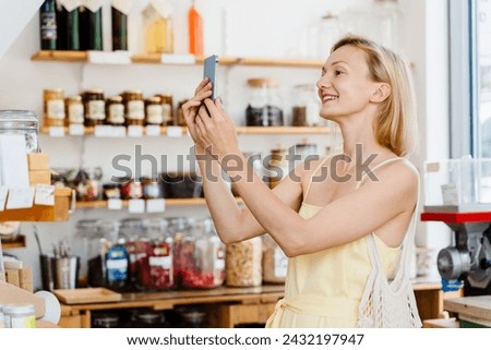 Smiling woman using smartphone and buying products at store. Сonscious shopping and retail of organic food and eco goods at zero waste shop. Happy girl taking photos at small local shop.