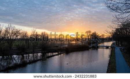 This serene image captures a sunset by a calm canal, with the sun dipping low in the sky and casting a vibrant orange hue along the horizon. The silhouette of leafless trees reflects on the water's