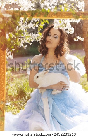 A beautiful girl in a blue dress with a decorative white rabbit against the background of a blooming apple tree. Animals and people.