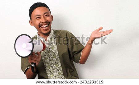 Excited Indonesian Muslim man in koko and peci gestures to the side with an open hand, holding a megaphone. He directs attention to empty copy space for text or advertisements. Isolated on a White