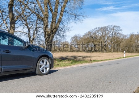 Anthracite colored front part of a car on the left side of the road with trees without leaves and a field in the background in bright sunshine and a blue sky