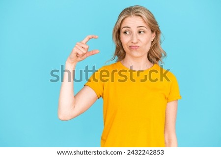 Young woman standing over blue background and showing small size gesture with hand. Sceptic female in yellow t-shirt doing sign little with fingers.