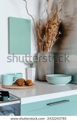 Detail in kitchen interior, cupboard with white countertop, clean blue plates, glass vase with dry plants, picture on wall and breakfast wooden tray with fresh croissants and ceramic cups
