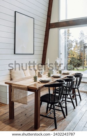 Setting table in dining room with modern interior, comfortable chairs, tablecloth runner, empty plates, wine glass, candles and picture in frame with copy space on wall. Dinner party preparation