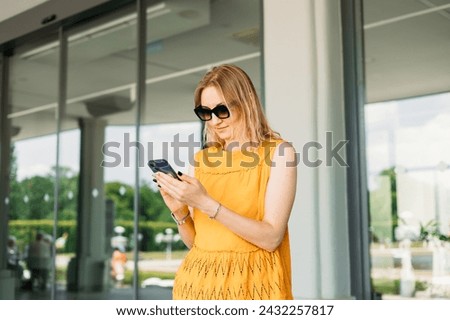 Happy cheerful young blond woman in sunglasses walking on city street checks her smartphone. Portrait of beautiful 30s girl using smartphone outdoors. Urban lifestyle concept. Summer time