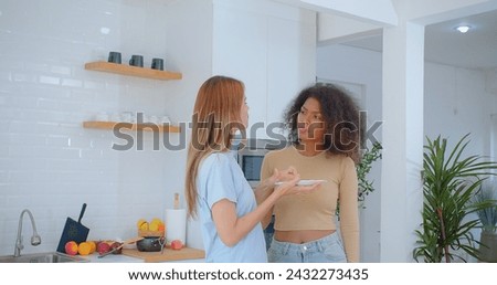 Laughter fills a sunlit kitchen as one woman presents a plate of food to her friend, who reacts with a delighted surprise