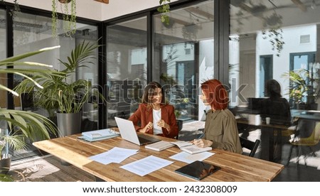Two busy business women of young and middle age talking in creative green office sitting at desk. Professional ladies employee and manager having conversation using laptop at work. Candid photo. Royalty-Free Stock Photo #2432308009