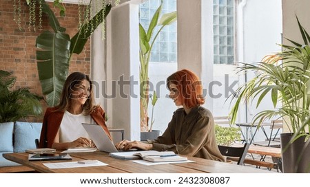 Two happy busy business women of young and middle age talking in green cozy office sitting at desk. Professional ladies executives having conversation using laptop at work. Authentic candid shot. Royalty-Free Stock Photo #2432308087
