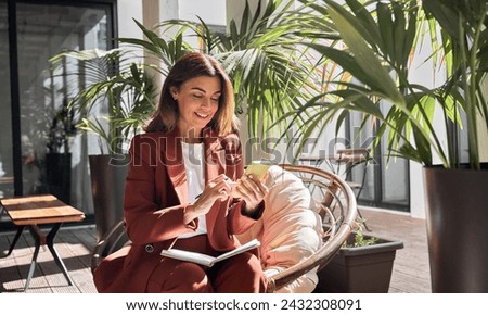 Happy smiling elegant mature middle aged business woman wearing suit holding cellphone using mobile cell phone looking at smartphone sitting in comfortable chair in sunny office with green plants.
