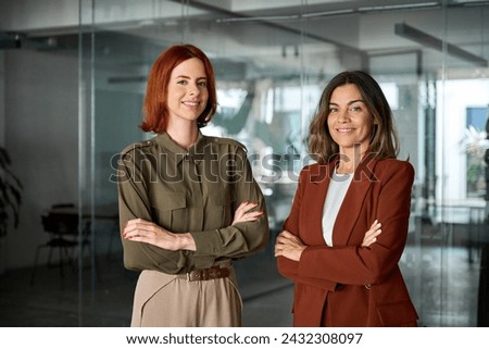 Happy confident professional business women of young and middle age posing at work looking at camera. Two smiling confident ladies standing arms crossed in office. Women power partnership, portrait. Royalty-Free Stock Photo #2432308097