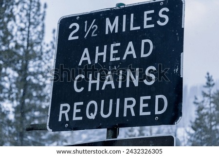 Highway sign in Sierra Nevada mountains that reads,"Two and a half Miles Ahead Chains Required"  Snowy winter backdrop with trees.  Captured during the onset of a winter storm.  